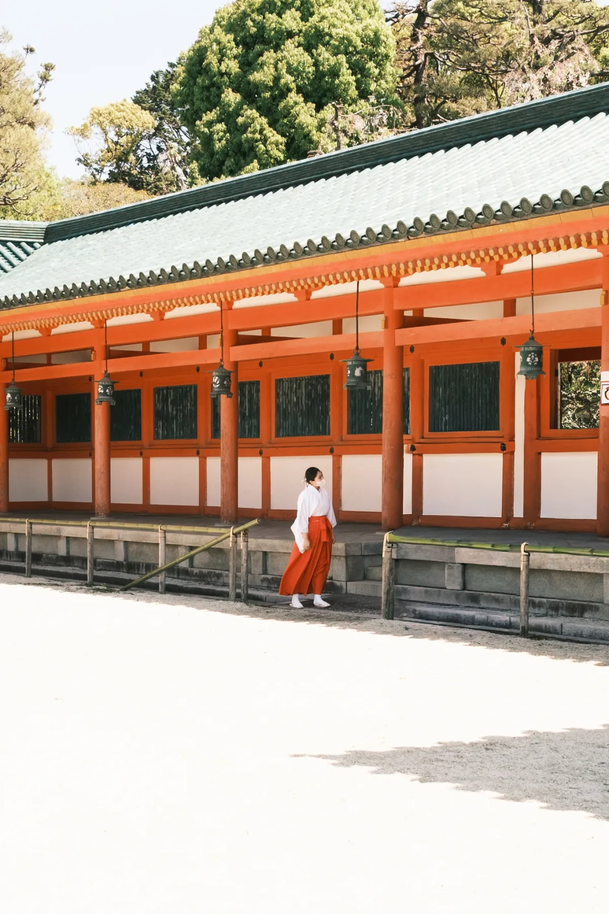 A shrine attendant in red and white walking past a long vermilion corridor.